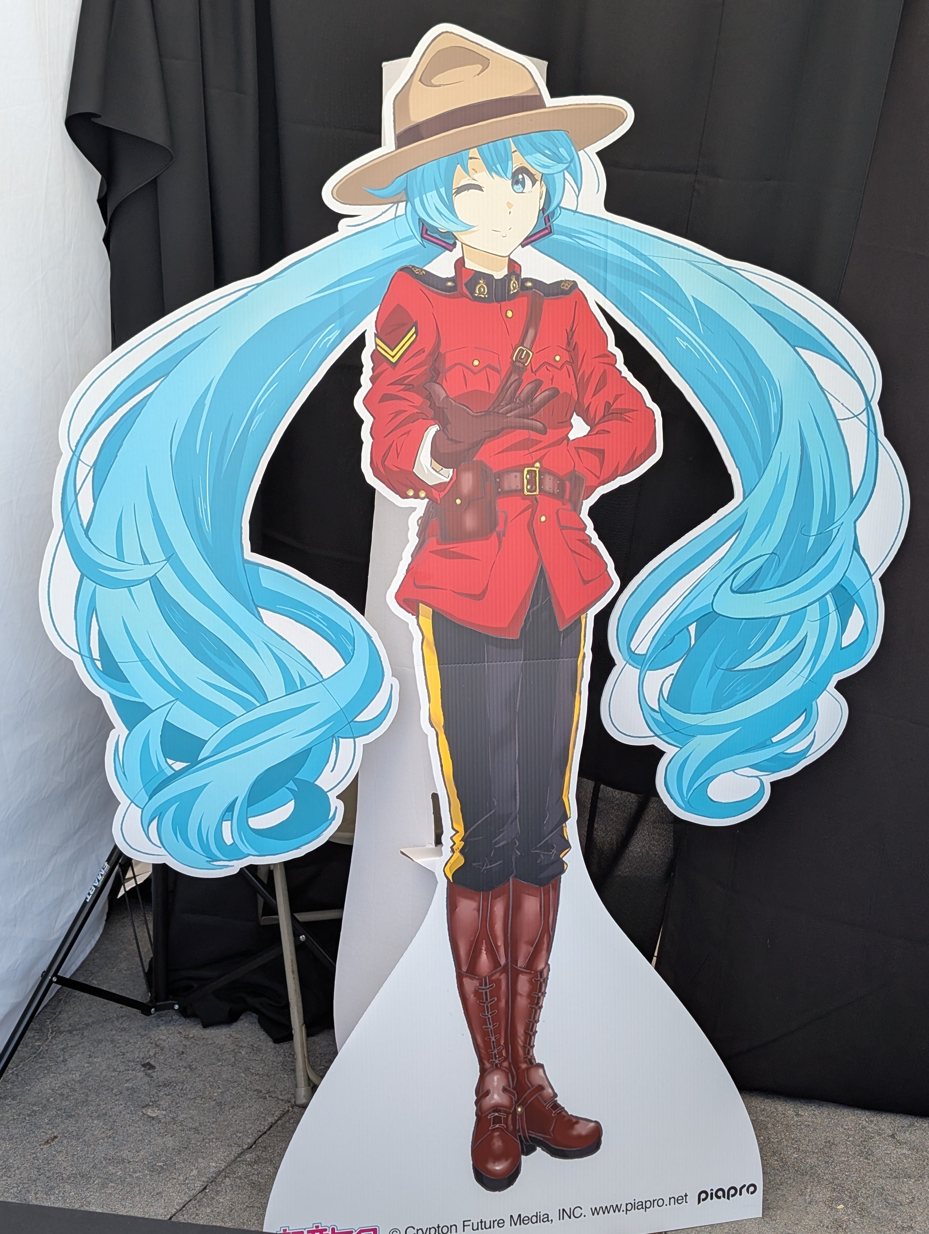 hatsune miku, royal canadian mounted police, vocaloid, absurdres, artist request, highres, official art, photo (medium), third-party source, 1girl, arm behind back, belt, belt buckle, belt pouch, between breasts, blue eyes, blue hair, boots, breasts, brown boots, brown hat, buckle, cardboard cutout, character name, closed mouth, coat, copyright name, dot nose, floating hair, full body, hair between eyes, hair ornament, hat, knee boots, long hair, looking at viewer, messy hair, military rank insignia, one eye closed, outdoors, outstretched hand, police, pouch, red coat, single vertical stripe, small breasts, smile, solo, standing, strap between breasts, very long hair