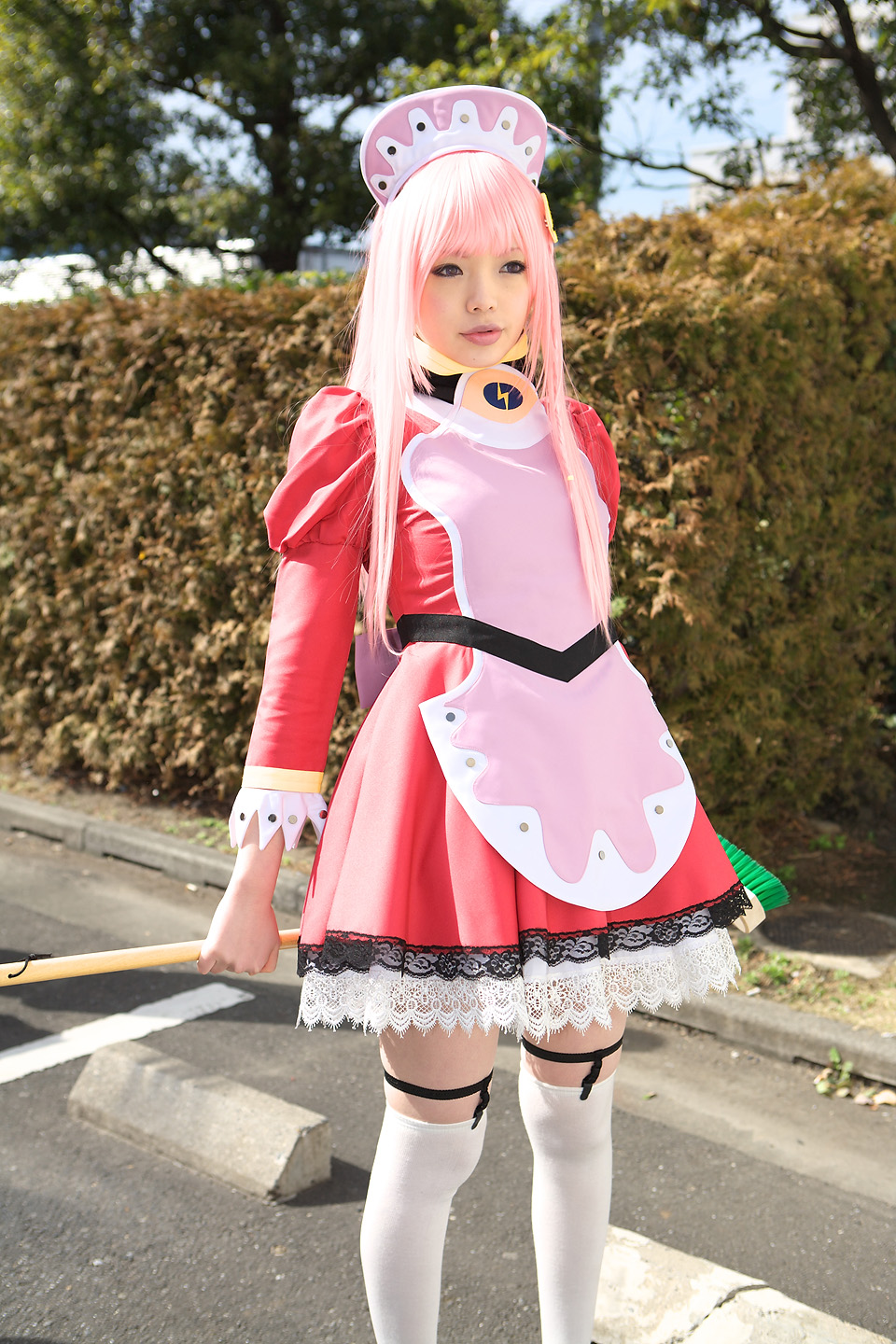 namada, nono (top wo nerae 2!), top wo nerae! (series), top wo nerae 2!, highres, photo (medium), 1girl, apron, asian, blurry, breasts, broom, cosplay, cowboy shot, depth of field, dress, female focus, garter straps, hedge, light smile, looking afar, maid, maid apron, makeup, mascara, outdoors, parking lot, parted lips, pink hair, puffy sleeves, short dress, sidelocks, small breasts, solo, standing, thighhighs, tree, white thighhighs, zettai ryouiki