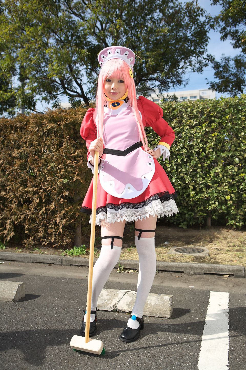 namada, nono (top wo nerae 2!), top wo nerae! (series), top wo nerae 2!, highres, photo (medium), 1girl, apron, asian, black shoes, blurry, breasts, broom, contrapposto, cosplay, depth of field, dress, female focus, garter straps, hand on own hip, hedge, light smile, looking at viewer, maid, maid apron, makeup, mary janes, mascara, outdoors, parking lot, pigeon-toed, pink hair, puffy sleeves, shoes, short dress, sidelocks, small breasts, solo, standing, thighhighs, tree, white thighhighs, zettai ryouiki