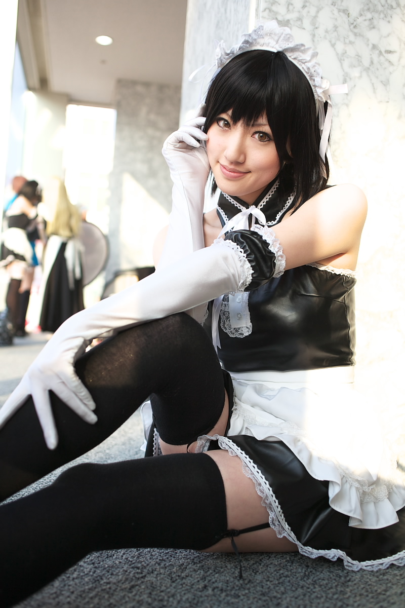 saya (cosplayer), sawatari izumi, kore ga watashi no goshujin-sama, highres, photo (medium), 1girl, apron, asian, bare shoulders, black hair, blurry, cosplay, cowboy shot, depth of field, detached collar, elbow gloves, facing viewer, feet out of frame, finger to mouth, garter straps, gloves, japanese (nationality), knee up, looking at viewer, maid, maid apron, maid headdress, nakabayashi yoshitaka's maid uniform, on floor, sawatari izumi (cosplay), short hair, sitting, solo focus, thighhighs, waist apron, white apron, white gloves