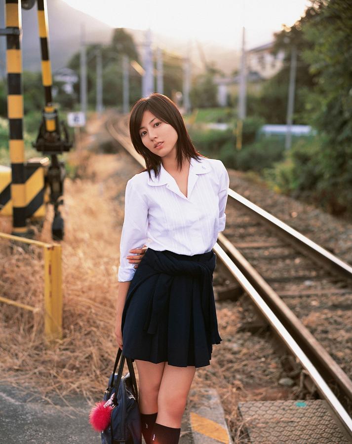 sugimoto yumi, photo (medium), 1girl, asian, bag, brown hair, female focus, kneehighs, long hair, looking at viewer, miniskirt, pleated skirt, railroad crossing, railroad tracks, school bag, school uniform, skirt, socks, solo, standing