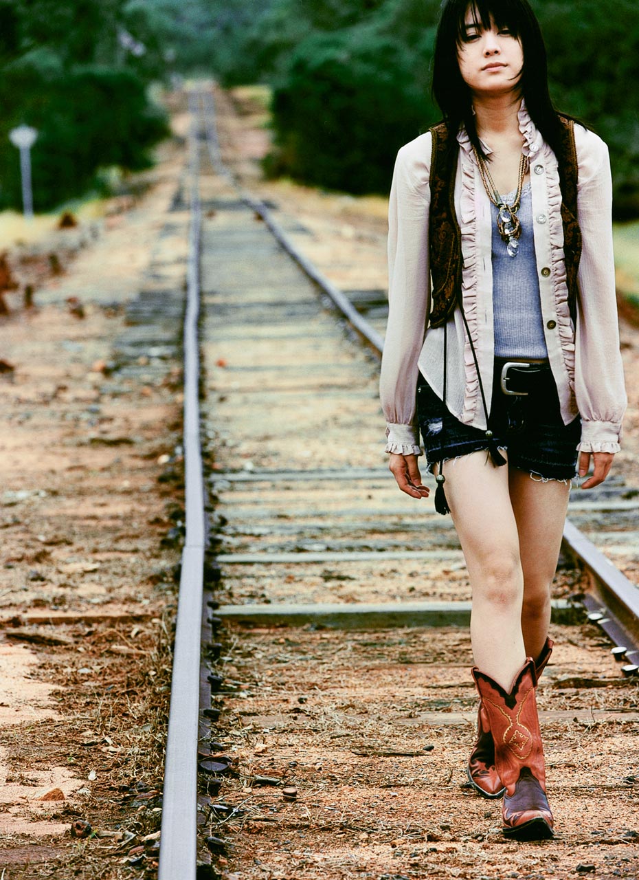 highres, photo (medium), 1girl, belt, black hair, boots, cowboy boots, female focus, fukada saki, jewelry, necklace, open clothes, open shirt, outdoors, railroad tracks, shirt, shorts, solo, tank top, vest, walking