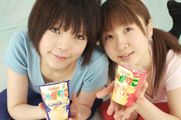 miasa, namada, black legwear, photo (medium), 2girls, asian, black hair, brown eyes, brown hair, color contacts, food, indoors, long hair, looking at viewer, lying, multiple girls, no shoes, on stomach, pillow, shirt, short hair, smile, snack, t-shirt, thighhighs, twintails