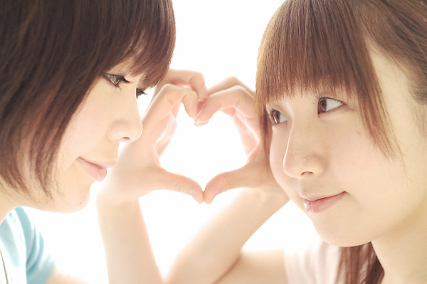 miasa, namada, photo (medium), translated, 2girls, asian, black hair, brown eyes, brown hair, eye contact, heart, heart hands, heart hands duo, indoors, long hair, looking at another, multiple girls, shirt, short hair, t-shirt, twintails