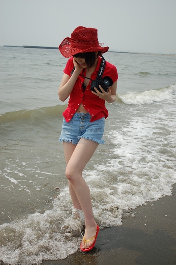 namada, photo (medium), 1girl, asian, bare legs, beach, black bra, black hair, bra, breasts, camera, cowboy hat, cutoffs, denim, denim shorts, female focus, flip-flops, full body, hat, lingerie, open clothes, open shirt, outdoors, red shirt, sandals, shirt, short hair, short sleeves, shorts, small breasts, solo, standing, underwear