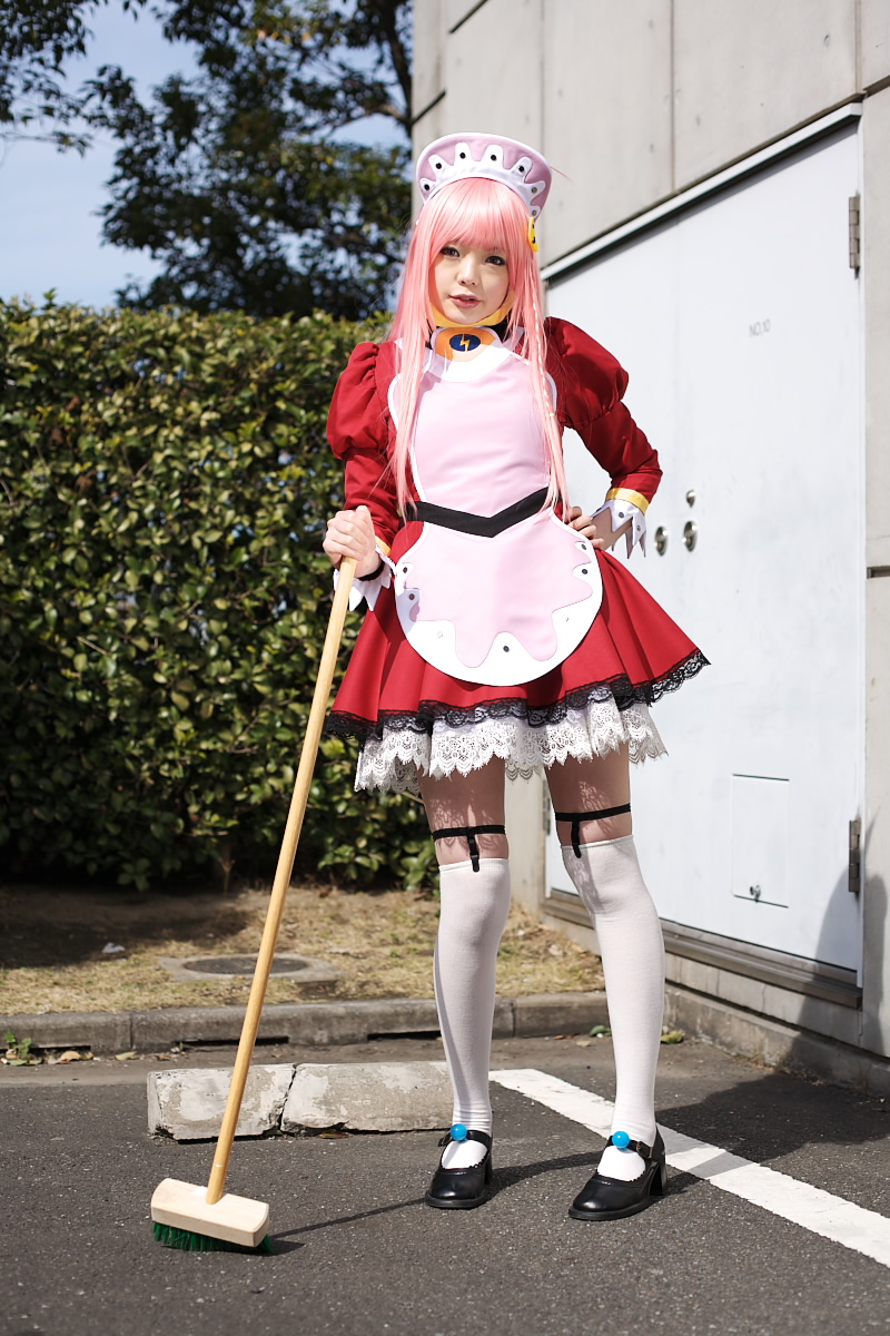 namada, nono (top wo nerae 2!), top wo nerae! (series), top wo nerae 2!, highres, photo (medium), 1girl, apron, asian, black shoes, blurry, breasts, broom, contrapposto, cosplay, depth of field, dress, female focus, garter straps, hand on own hip, hedge, looking at viewer, maid, maid apron, makeup, mary janes, mascara, outdoors, parking lot, parted lips, pigeon-toed, pink hair, puffy sleeves, shoes, short dress, sidelocks, small breasts, solo, standing, thighhighs, tree, white thighhighs, zettai ryouiki