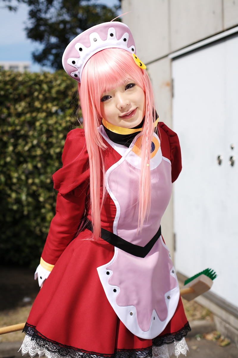 namada, nono (top wo nerae 2!), top wo nerae! (series), top wo nerae 2!, highres, photo (medium), 1girl, apron, asian, blurry, breasts, broom, cosplay, cowboy shot, depth of field, dress, female focus, head tilt, hedge, light smile, looking at viewer, maid, maid apron, makeup, mascara, outdoors, pink hair, puffy sleeves, sidelocks, small breasts, solo, standing