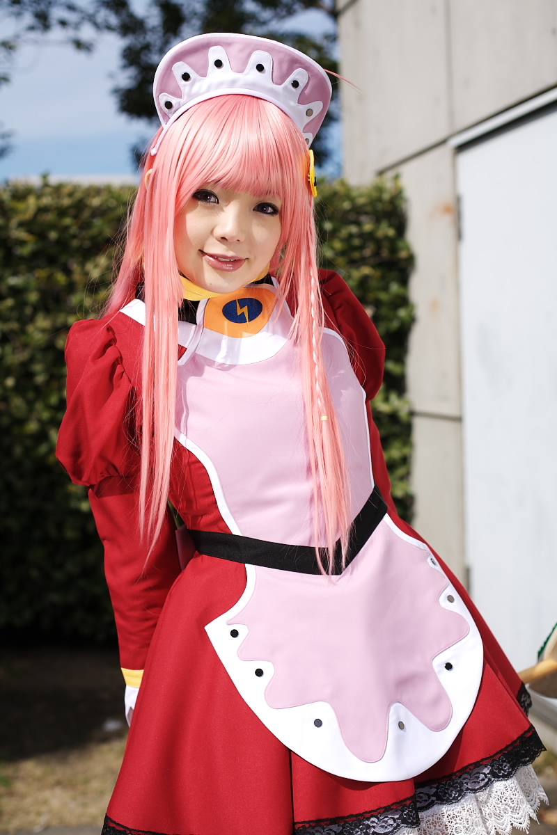 namada, nono (top wo nerae 2!), top wo nerae! (series), top wo nerae 2!, highres, photo (medium), 1girl, apron, asian, blurry, breasts, broom, contrapposto, cosplay, cowboy shot, depth of field, dress, female focus, hedge, light smile, looking at viewer, maid, maid apron, makeup, mascara, outdoors, pink hair, puffy sleeves, sidelocks, small breasts, solo, standing
