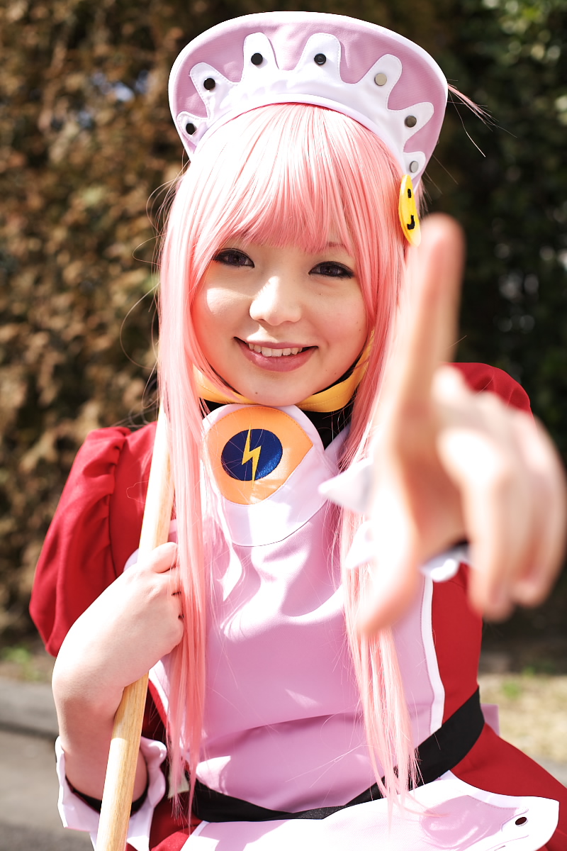 namada, nono (top wo nerae 2!), top wo nerae! (series), top wo nerae 2!, highres, photo (medium), 1girl, apron, asian, blurry, breasts, broom, cosplay, depth of field, dress, female focus, hedge, index finger raised, looking at viewer, maid, maid apron, makeup, mascara, outdoors, parking lot, pink hair, pointing, pointing at viewer, puffy sleeves, sidelocks, small breasts, smile, solo