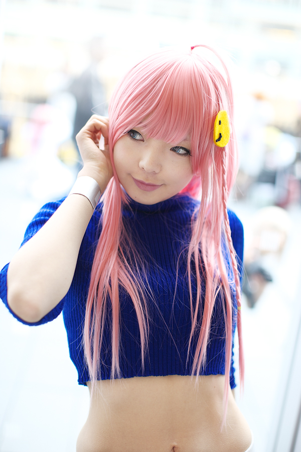 namada, nono (top wo nerae 2!), top wo nerae! (series), top wo nerae 2!, photo (medium), 1girl, adjusting hair, asian, blue eyes, blurry, braid, breasts, color contacts, cosplay, depth of field, female focus, head tilt, indoors, light smile, looking to the side, makeup, mascara, midriff, pink hair, side braid, sidelocks, small breasts, solo, standing, sweater