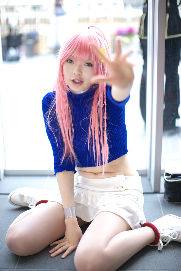 namada, nono (top wo nerae 2!), top wo nerae! (series), top wo nerae 2!, photo (medium), 1girl, arm support, asian, blue eyes, blue sweater, blurry, braid, breasts, color contacts, cosplay, depth of field, female focus, frilled skirt, frills, indoors, long hair, looking at viewer, makeup, mascara, midriff, miniskirt, navel, outstretched arm, pink hair, side braid, sidelocks, sitting, skirt, small breasts, solo, sweater, wariza, white skirt