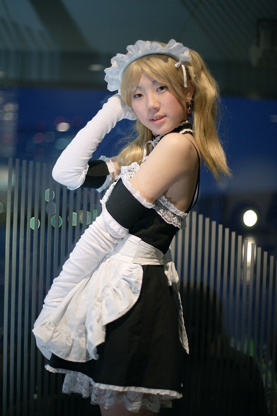 hime (cosplayer), sawatari izumi, kore ga watashi no goshujin-sama, highres, photo (medium), 1girl, apron, asian, bare shoulders, blonde hair, blurry, cosplay, cowboy shot, depth of field, detached collar, earrings, elbow gloves, female focus, gloves, indoors, japanese (nationality), jewelry, looking at viewer, maid, maid apron, maid headdress, nakabayashi yoshitaka's maid uniform, night, sawatari izumi (cosplay), solo, twintails, white gloves