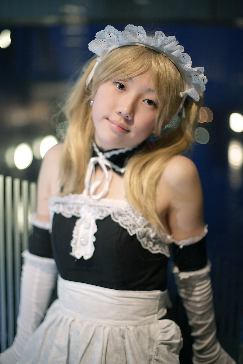 hime (cosplayer), sawatari izumi, kore ga watashi no goshujin-sama, highres, photo (medium), 1girl, apron, asian, bare shoulders, blonde hair, blurry, cosplay, depth of field, detached collar, earrings, elbow gloves, female focus, gloves, indoors, japanese (nationality), jewelry, looking at viewer, maid, maid apron, maid headdress, nakabayashi yoshitaka's maid uniform, sawatari izumi (cosplay), solo, twintails, upper body, white gloves