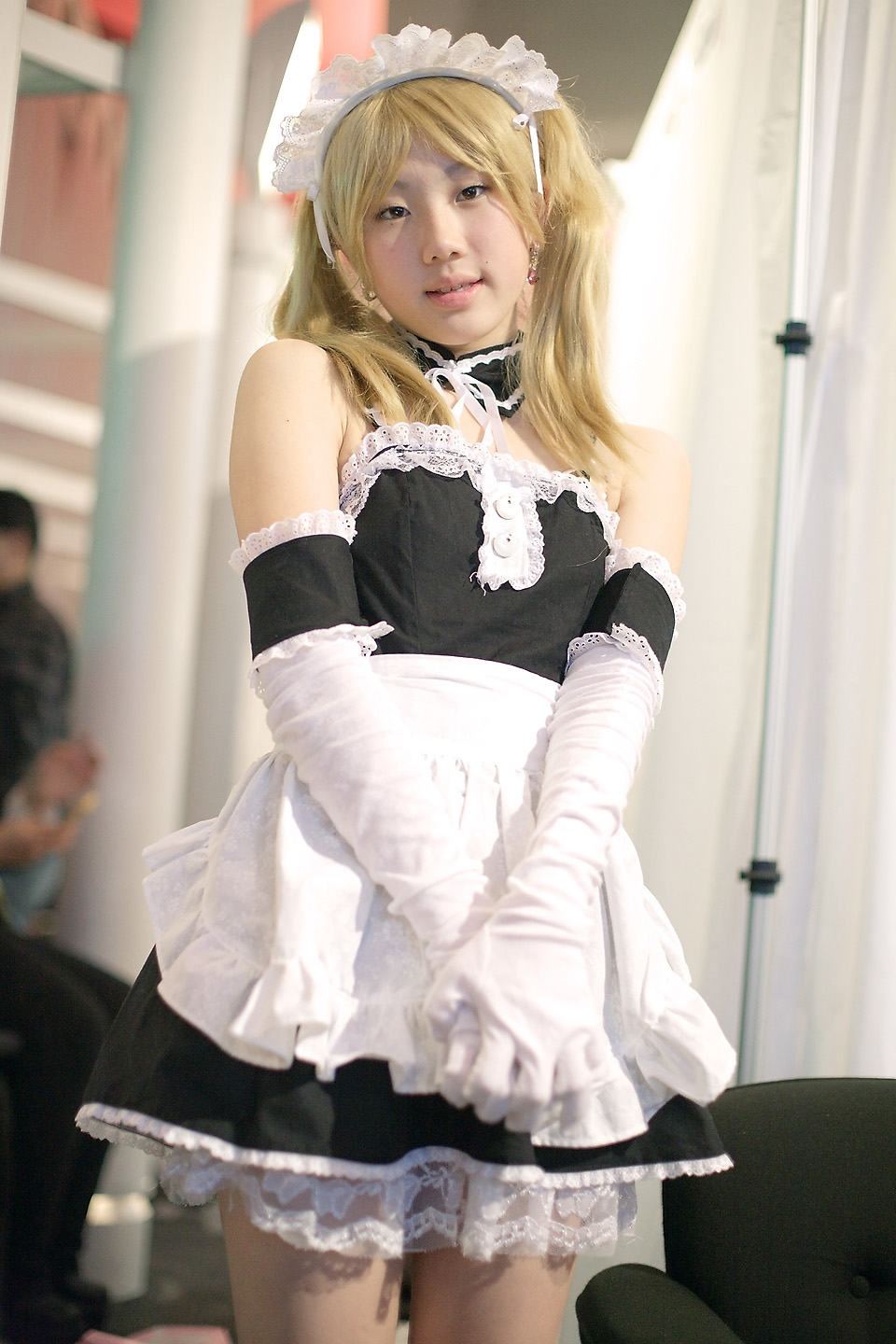 hime (cosplayer), sawatari izumi, kore ga watashi no goshujin-sama, highres, photo (medium), 1girl, apron, asian, bare shoulders, blonde hair, blurry, cosplay, cowboy shot, depth of field, detached collar, earrings, elbow gloves, female focus, gloves, indoors, japanese (nationality), jewelry, looking at viewer, maid, maid apron, maid headdress, nakabayashi yoshitaka's maid uniform, sawatari izumi (cosplay), solo, twintails, white gloves