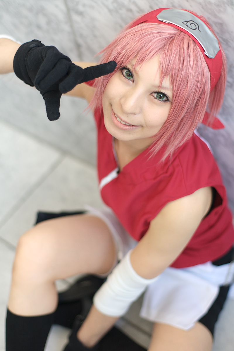 ari, haruno sakura, naruto (series), naruto shippuuden, black legwear, highres, photo (medium), 1girl, arm warmers, bare shoulders, black gloves, blurry, cosplay, depth of field, eyelashes, female focus, from above, gloves, green eyes, hand up, haruno sakura (cosplay), head tilt, headband, headdress, konohagakure symbol, lips, looking at viewer, looking up, miniskirt, parted lips, pink hair, red shirt, shirt, short hair, shorts, sitting, skirt, sleeveless, sleeveless shirt, smile, solo, white skirt