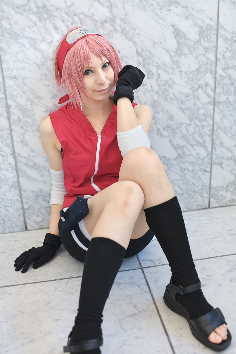 ari, haruno sakura, naruto (series), naruto shippuuden, highres, photo (medium), 1girl, arm warmers, bandages, bare shoulders, bike shorts, black gloves, black shoes, black socks, blurry, cosplay, depth of field, eyelashes, female focus, gloves, green eyes, hand up, haruno sakura (cosplay), head tilt, headband, headdress, holster, konohagakure symbol, lips, looking at viewer, on floor, pink hair, red shirt, shirt, shoes, short hair, shorts, sitting, skin tight, sleeveless, sleeveless shirt, smile, socks, solo, spandex, thigh holster, thigh strap, toeless legwear, toes