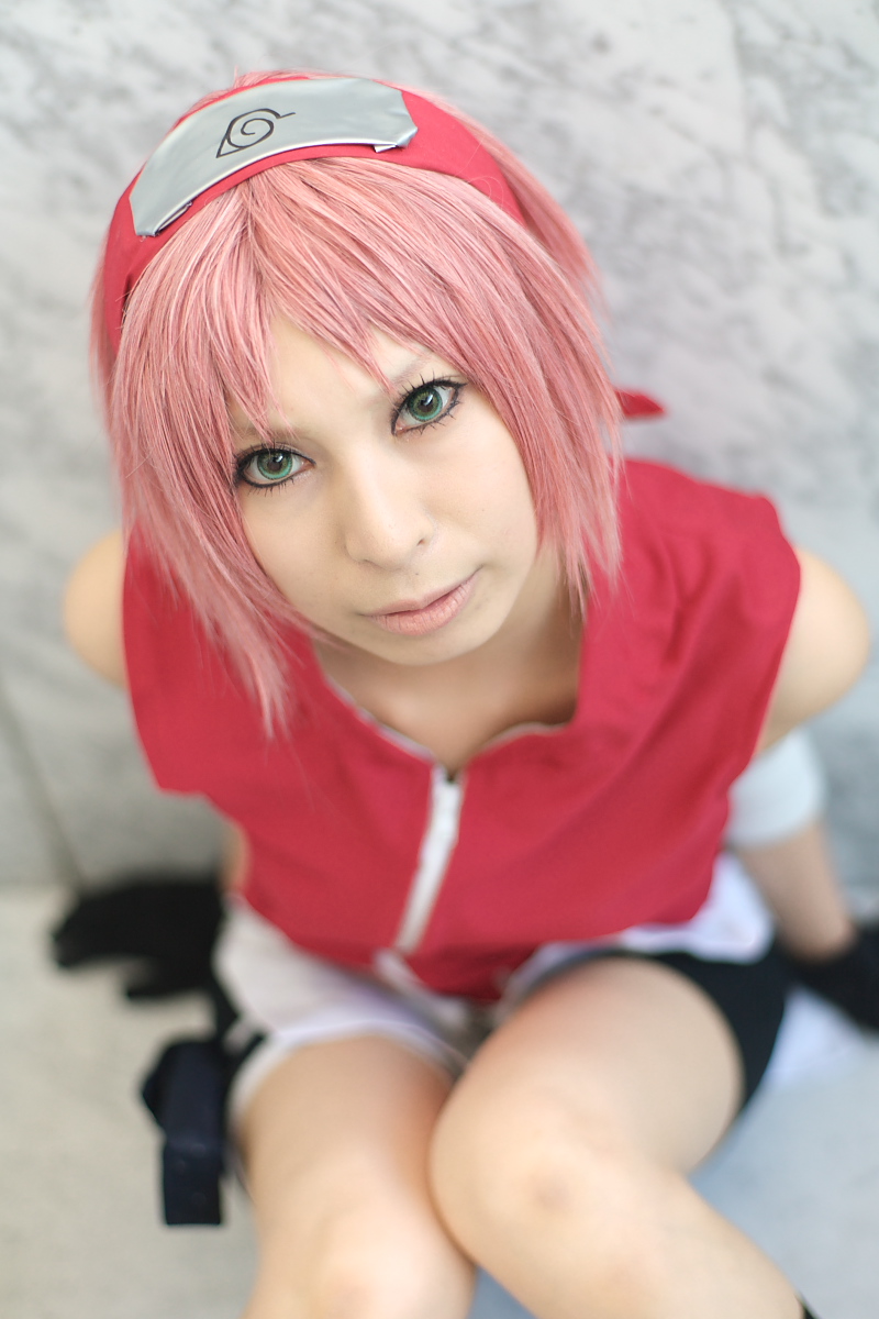 ari, naruto (series), naruto shippuuden, highres, photo (medium), 1girl, arm warmers, bandages, bare shoulders, black gloves, blurry, cosplay, depth of field, eyelashes, female focus, gloves, green eyes, haruno sakura (cosplay), head tilt, headband, headdress, holster, konohagakure symbol, lips, looking at viewer, looking up, pink hair, red shirt, shirt, short hair, shorts, side slit, sitting, skirt, sleeveless, sleeveless shirt, solo, thigh holster, thigh strap, white skirt