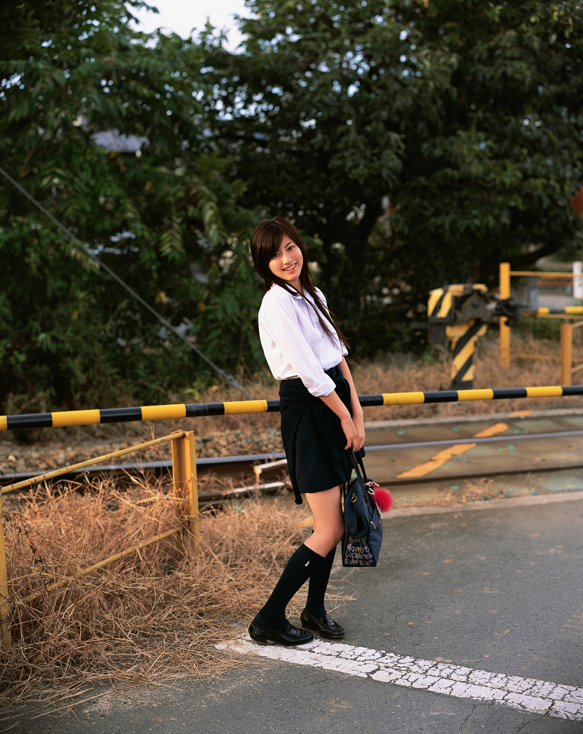 sugimoto yumi, highres, photo (medium), 1girl, asian, bag, brown hair, cosplay, female focus, kneehighs, long hair, looking at viewer, miniskirt, pleated skirt, railroad crossing, railroad tracks, school bag, school uniform, skirt, socks, solo, standing