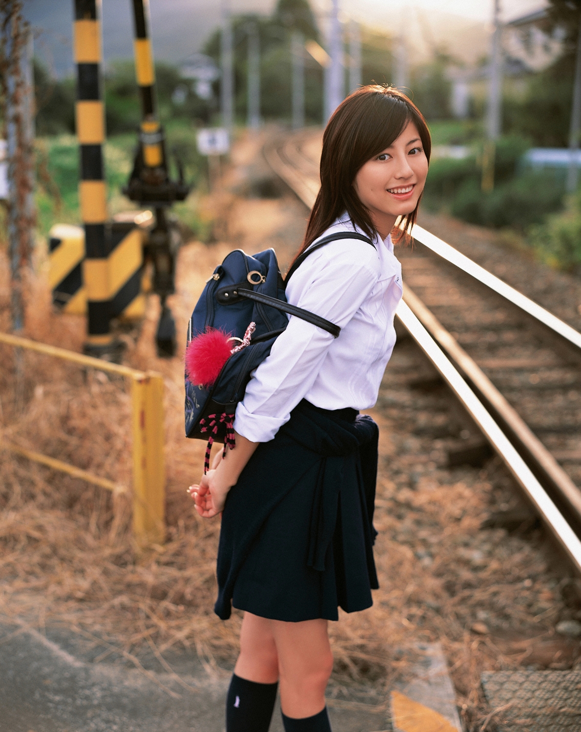 sugimoto yumi, highres, photo (medium), 1girl, asian, bag, brown hair, cosplay, female focus, kneehighs, long hair, looking at viewer, miniskirt, pleated skirt, railroad crossing, railroad tracks, school bag, school uniform, skirt, socks, solo, standing