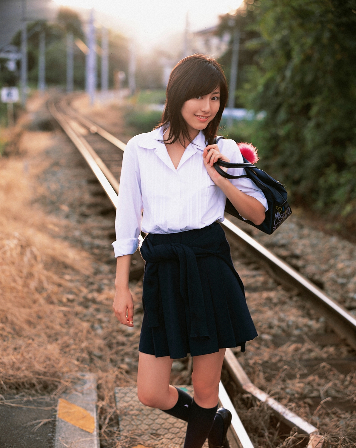 sugimoto yumi, highres, photo (medium), 1girl, asian, bag, brown hair, cosplay, female focus, kneehighs, long hair, looking at viewer, miniskirt, pleated skirt, railroad crossing, railroad tracks, school bag, school uniform, skirt, socks, solo, standing