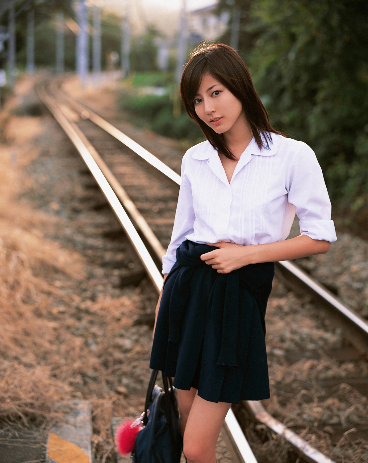 sugimoto yumi, highres, photo (medium), 1girl, asian, bag, brown hair, cosplay, female focus, kneehighs, long hair, looking at viewer, miniskirt, pleated skirt, railroad crossing, railroad tracks, school bag, school uniform, skirt, socks, solo, standing