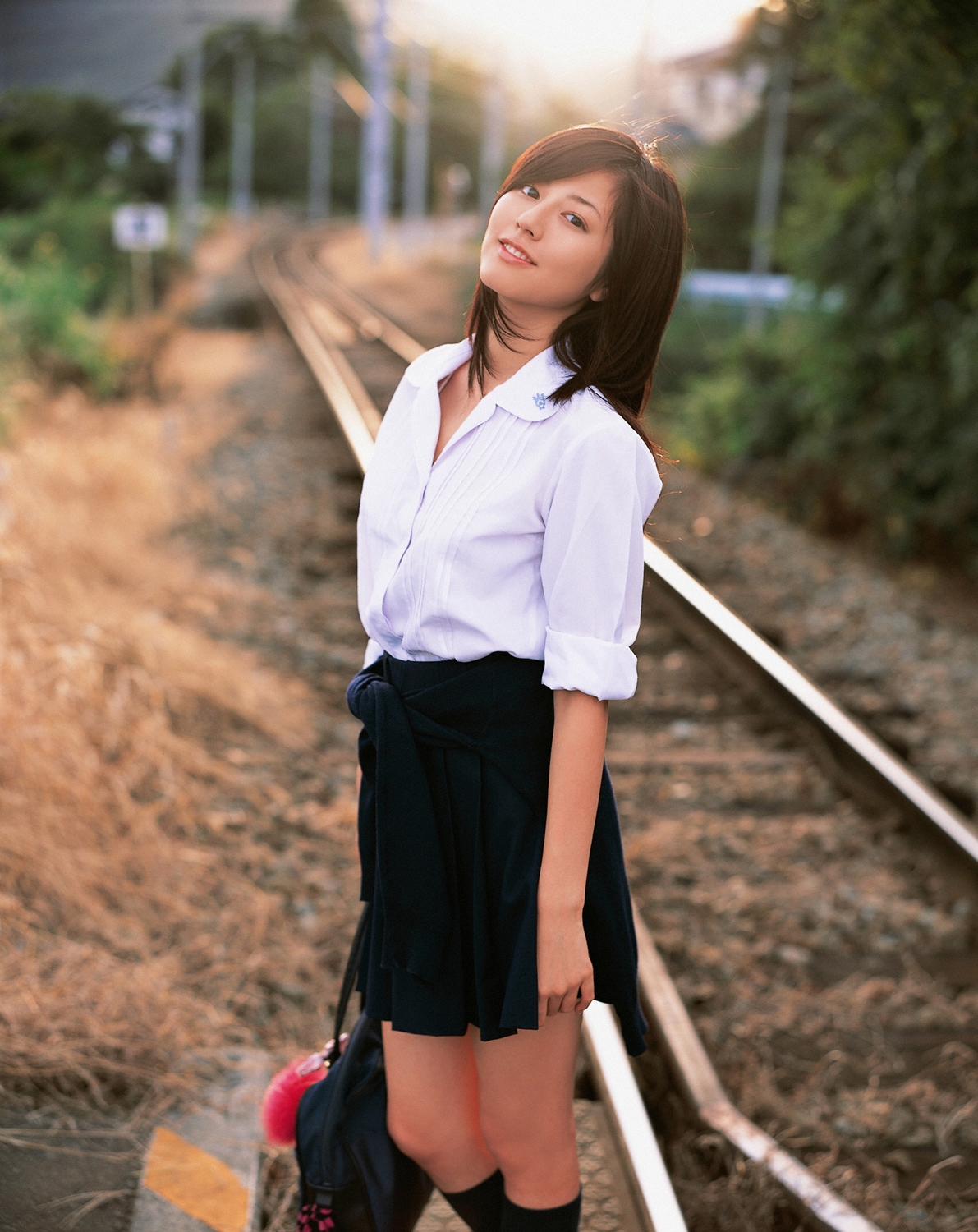 sugimoto yumi, highres, photo (medium), 1girl, asian, bag, brown hair, cosplay, female focus, kneehighs, long hair, looking at viewer, miniskirt, pleated skirt, railroad crossing, railroad tracks, school bag, school uniform, skirt, socks, solo, standing