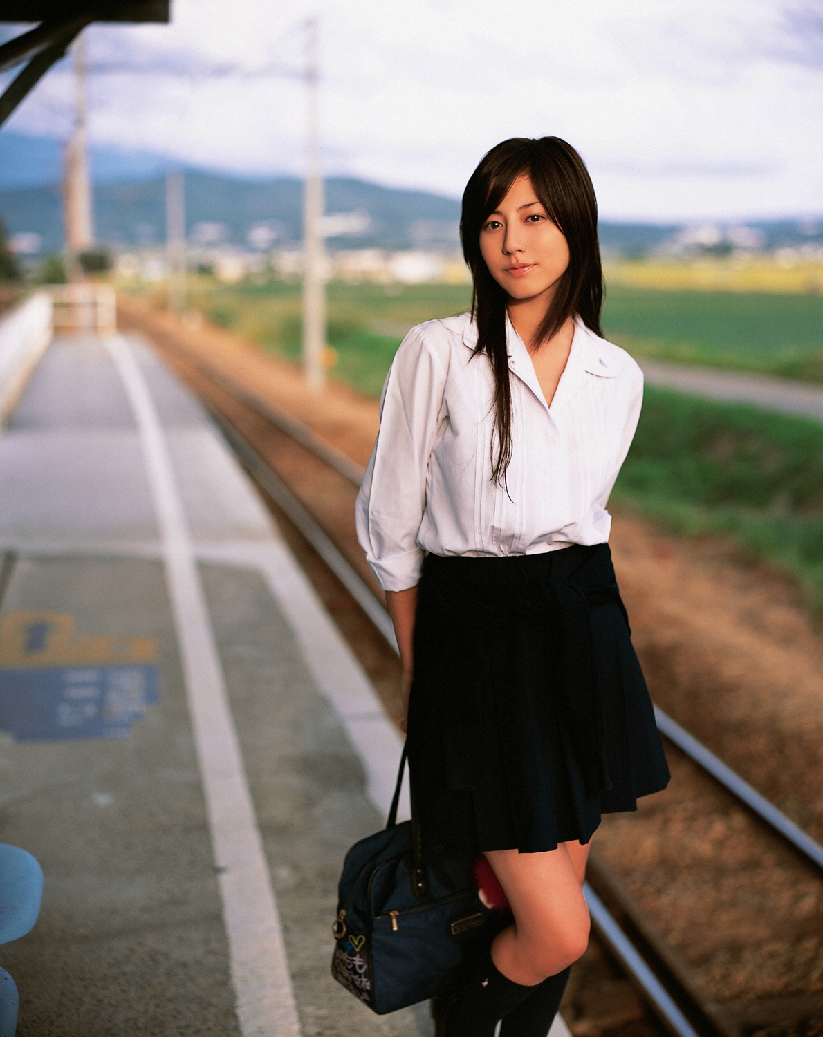 sugimoto yumi, highres, photo (medium), 1girl, asian, bag, brown hair, cosplay, female focus, kneehighs, long hair, looking at viewer, miniskirt, pleated skirt, railroad tracks, school bag, school uniform, skirt, socks, solo, standing, train station