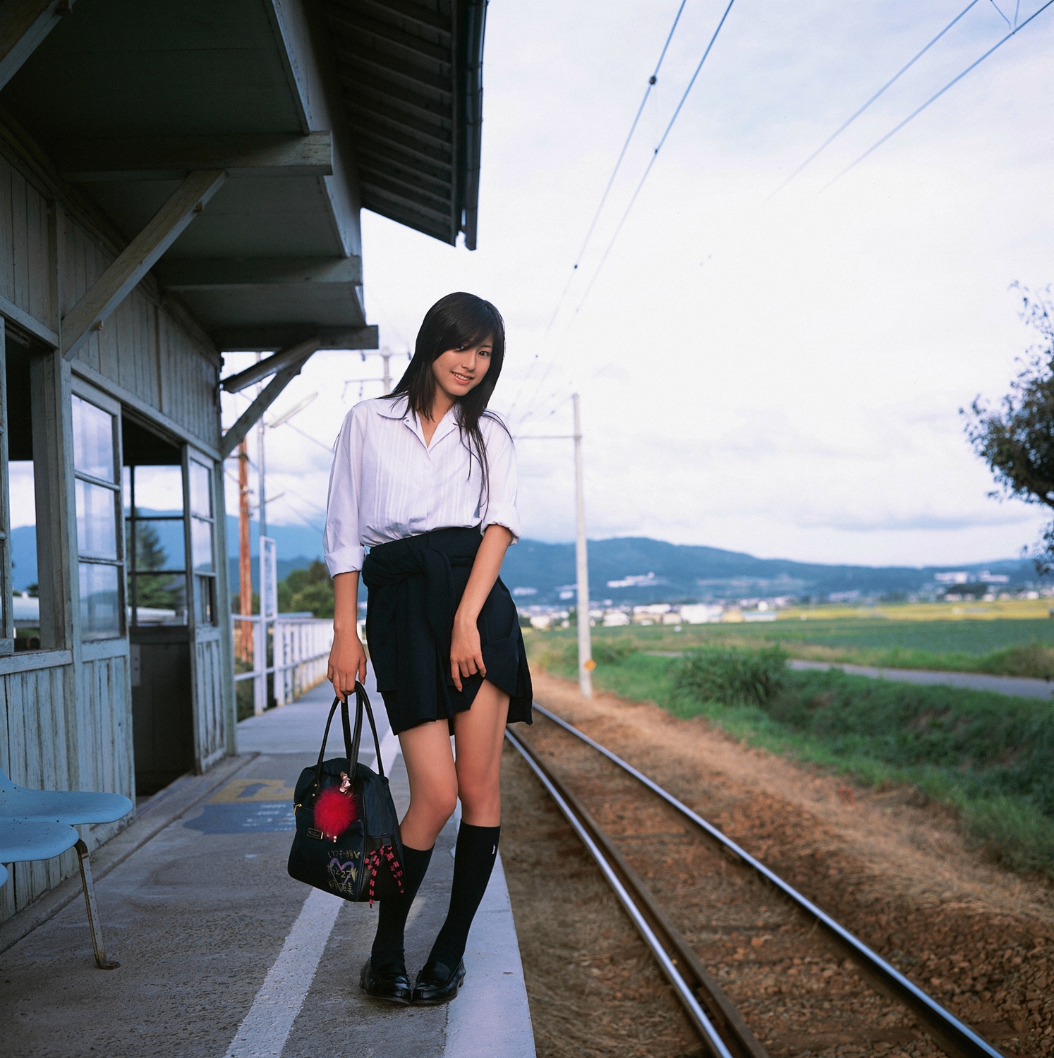 sugimoto yumi, highres, photo (medium), 1girl, asian, bag, brown hair, clothes lift, cosplay, female focus, kneehighs, long hair, looking at viewer, miniskirt, pleated skirt, railroad tracks, school bag, school uniform, skirt, skirt lift, socks, solo, standing, train station