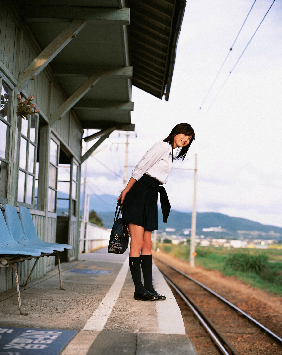 sugimoto yumi, highres, photo (medium), 1girl, asian, bag, brown hair, cosplay, female focus, kneehighs, long hair, looking at viewer, miniskirt, pleated skirt, railroad tracks, school bag, school uniform, skirt, socks, solo, standing, train station