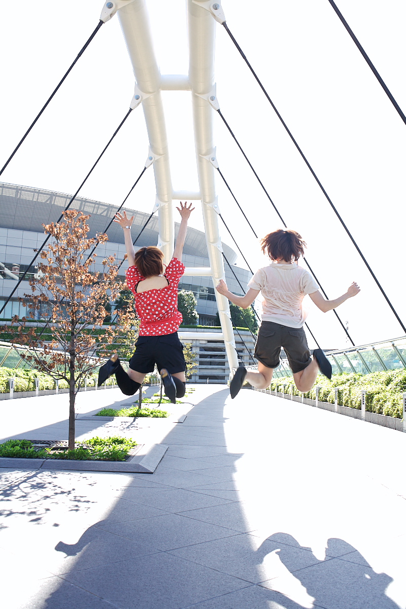 ari (model), chippi, highres, photo (medium), 2girls, asian, black shorts, bridge, brown hair, building, cosplay, dyed hair, female focus, from behind, jumping, multiple girls, outdoors, plant, polka dot, polka dot shirt, shadow, shirt, short hair, shorts, thighhighs, tree