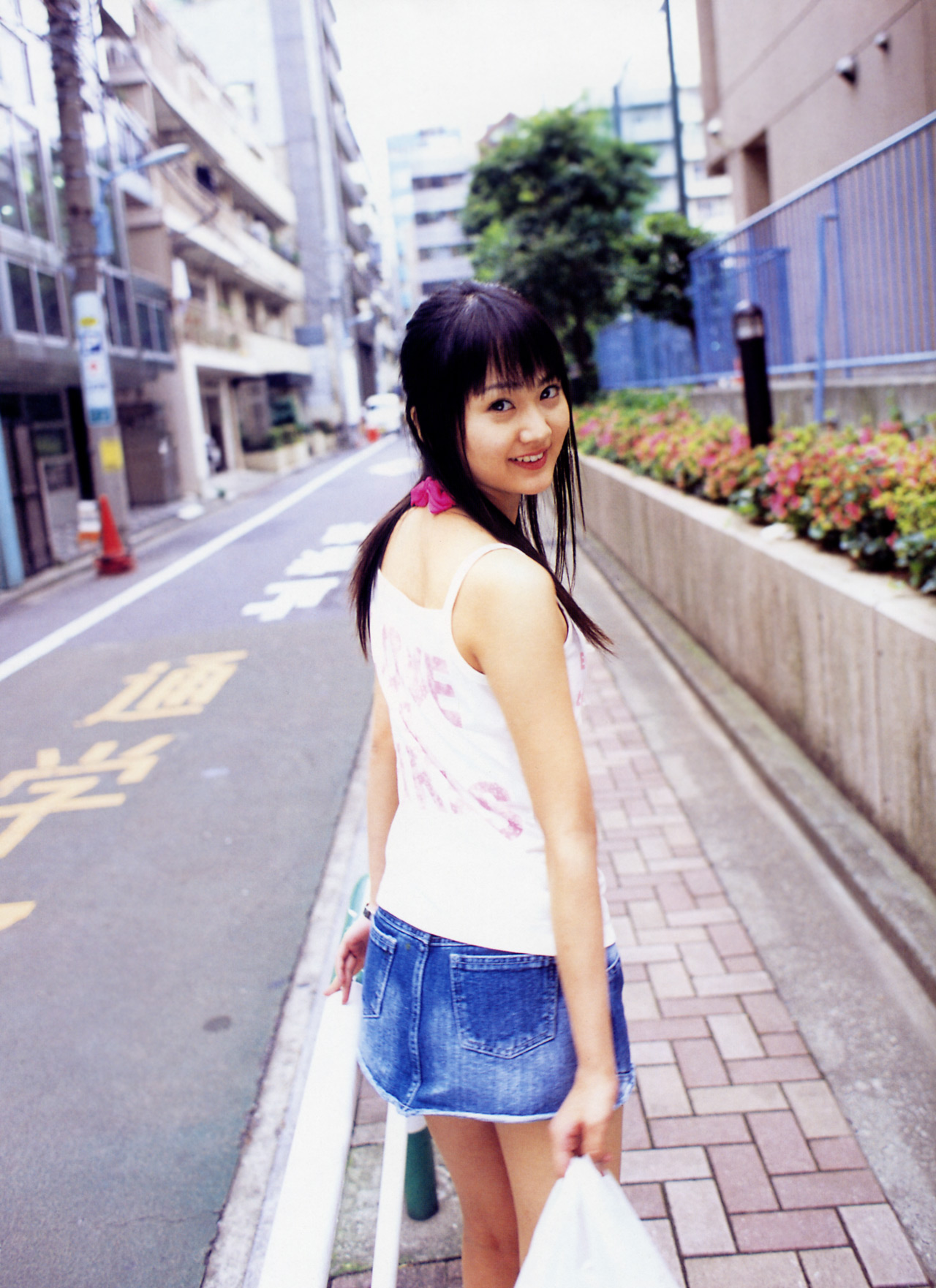 hamada shoko, lolita race queen, highres, photo (medium), 1girl, asian, bag, bare legs, black hair, breasts, denim, denim skirt, female focus, from behind, long hair, looking at viewer, looking back, miniskirt, outdoors, road, shopping bag, skirt, small breasts, smile, solo, street, tank top