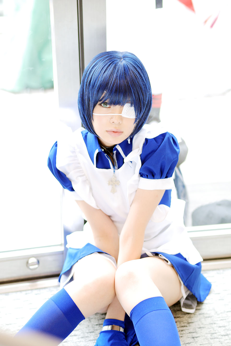 namada, ryomou shimei, ikkitousen, highres, photo (medium), 1girl, apron, arms between legs, asian, blue eyes, blue gloves, blue hair, blue socks, collar, color contacts, cosplay, cross, eyepatch, female focus, gloves, indoors, kneehighs, knees together feet apart, looking at viewer, maid, maid apron, on floor, puffy short sleeves, puffy sleeves, ryomou shimei (cosplay), short sleeves, sitting, socks, solo