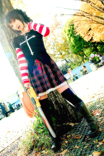 omi gibson, frederica sawyer, black lagoon, black footwear, striped, photo (medium), 1girl, asian, boots, chainsaw, cosplay, cross, cross print, dutch angle, electrolarynx, female focus, frederica sawyer (cosplay), full body, looking at viewer, outdoors, pleated skirt, skirt, solo focus, standing, thighhighs, tree