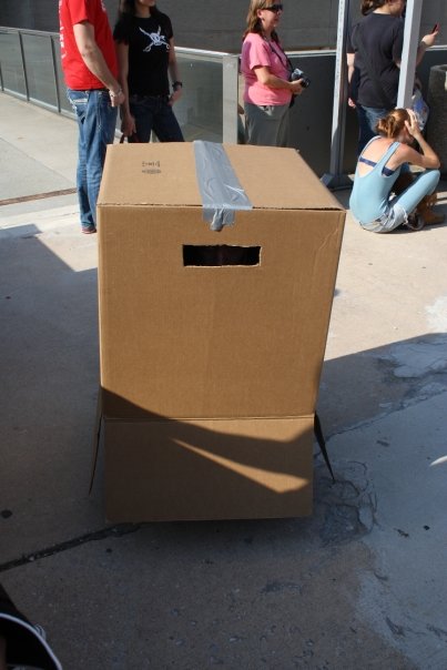 solid snake, metal gear (series), metal gear solid, brown footwear, cosplay photo, photo (medium), self-upload, bare arms, bare shoulders, black shirt, blue bra, boots, box, bra, camera, cardboard box, cosplay, day, denim, jeans, knee boots, outdoors, pants, peeking out, pink shirt, ponytail, red shirt, shirt, short sleeves, shorts, sitting, skull print, standing, sunglasses, sunlight, underwear, white shorts